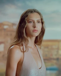 Portrait of a female model wearing a pale halter dress against an atmospheric painted backdrop with distant architecture.
