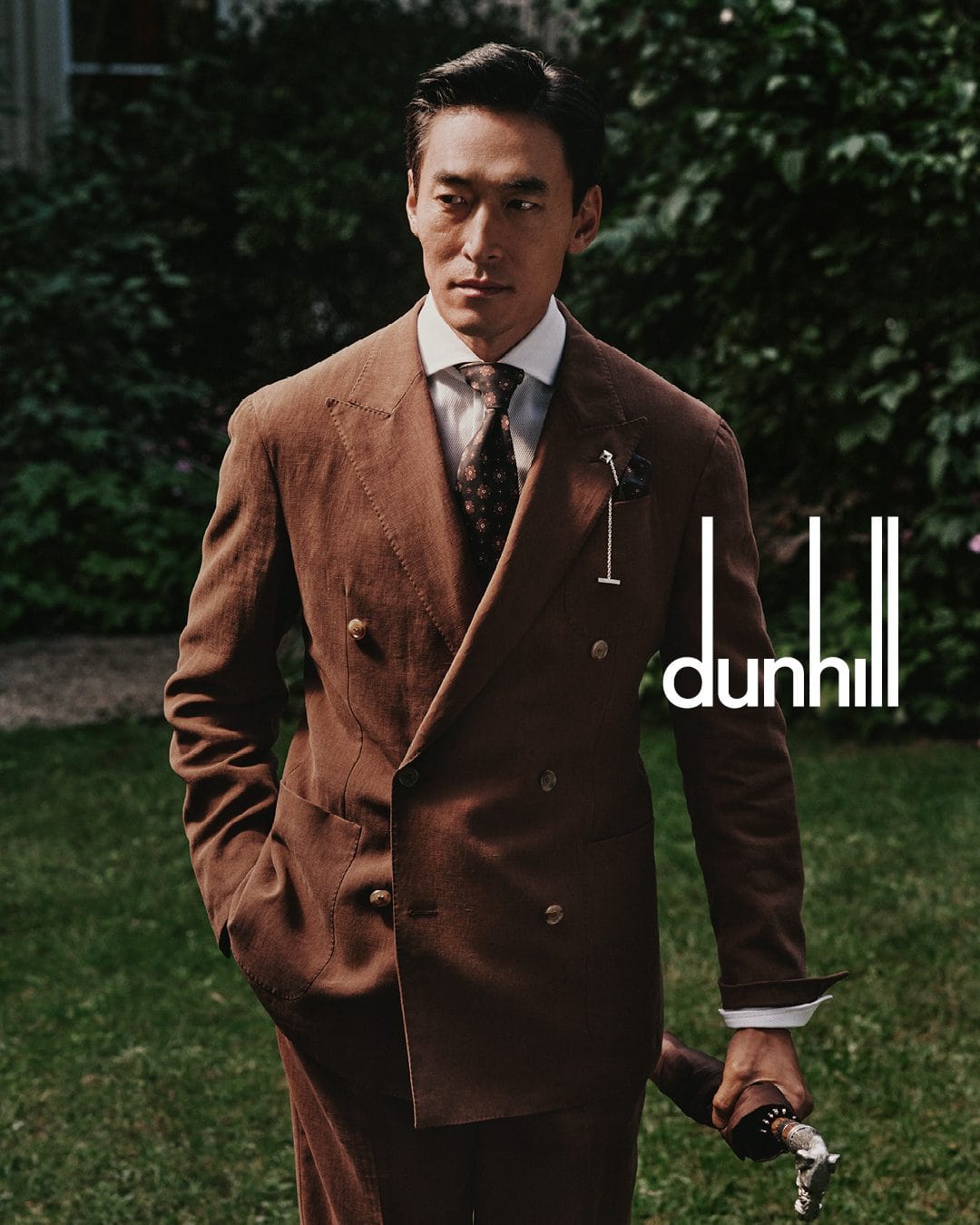 Asian man in a brown double-breasted suit standing outdoors in a garden for the Dunhill Spring 2025 ad campaign
