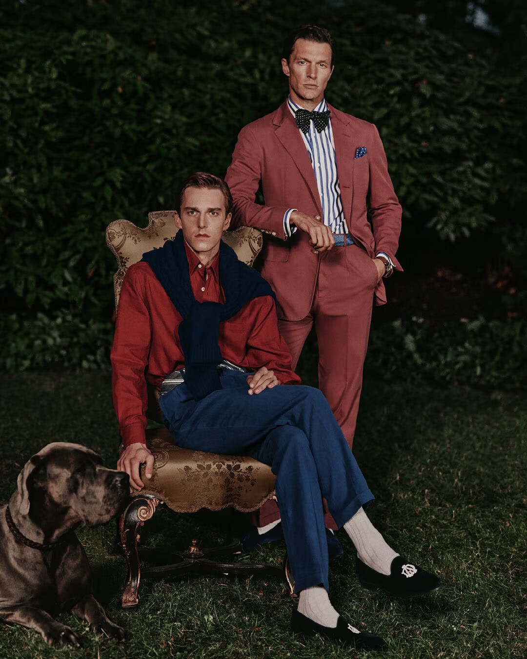 Two men posing outdoors for Dunhill Spring 2025 ad campaign, one seated on an ornate chair with a large dog, one standing behind.
