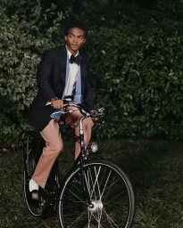 A young Black man wearing a tailored blazer and bow tie riding a bicycle outdoors against a lush green foliage background.