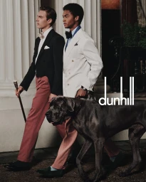 Two men in formalwear walking alongside a large dark dog near classical architectural columns for Dunhill Spring 2025 campaign.