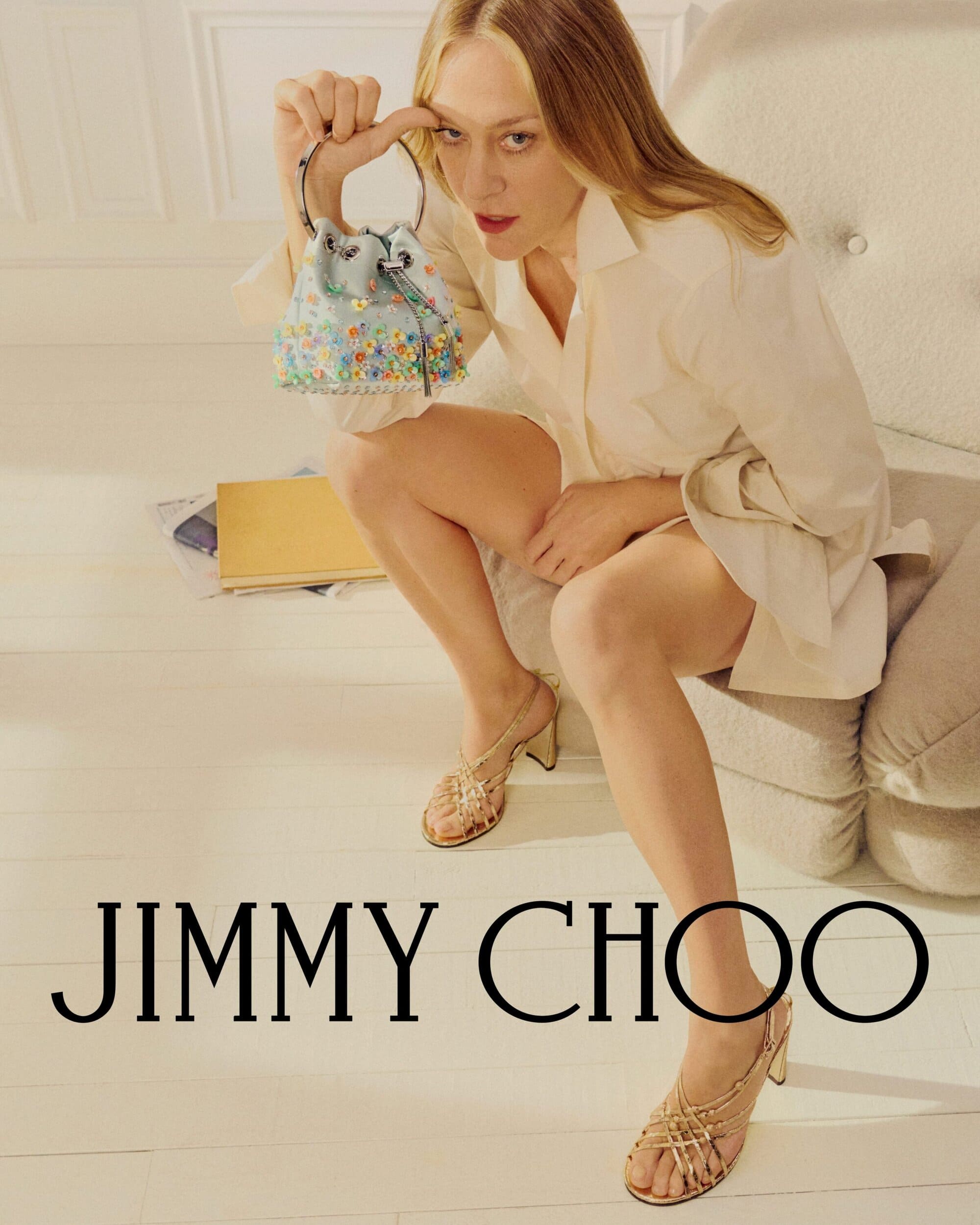 Chloe Sevigny seated on a white chair holding an embellished bucket bag for Jimmy Choo summer campaign