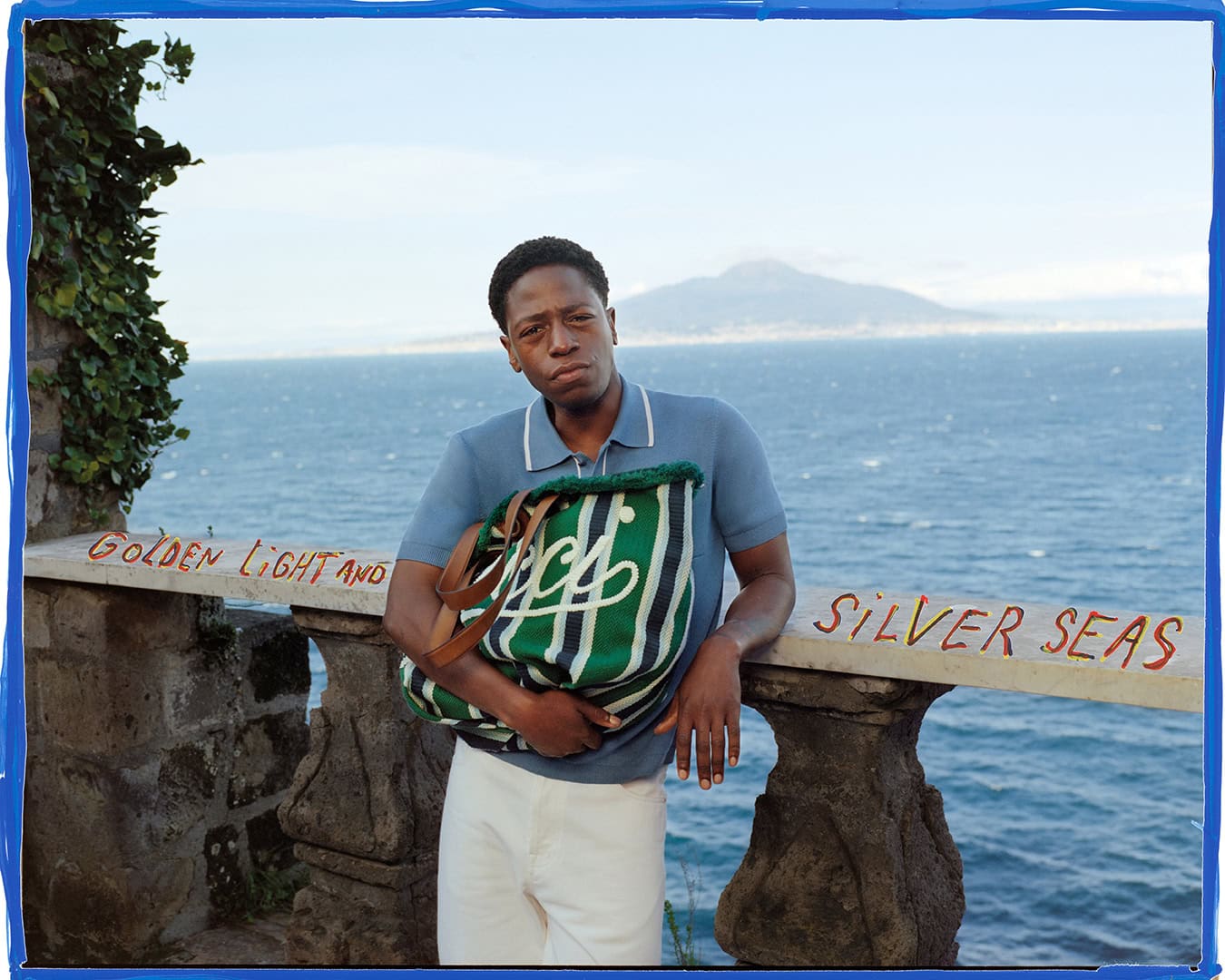 Man wearing a blue polo shirt and white trousers leaning on a stone balustrade holding a striped green and white tote bag with the ocean in the background
