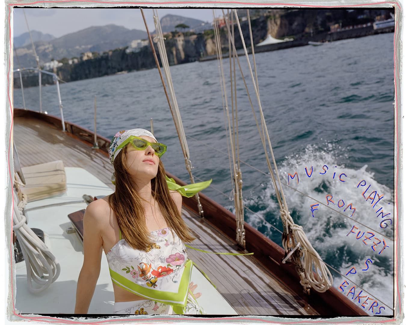 Woman wearing floral print top, green sunglasses, and headscarf seated on a wooden sailboat deck with ocean background