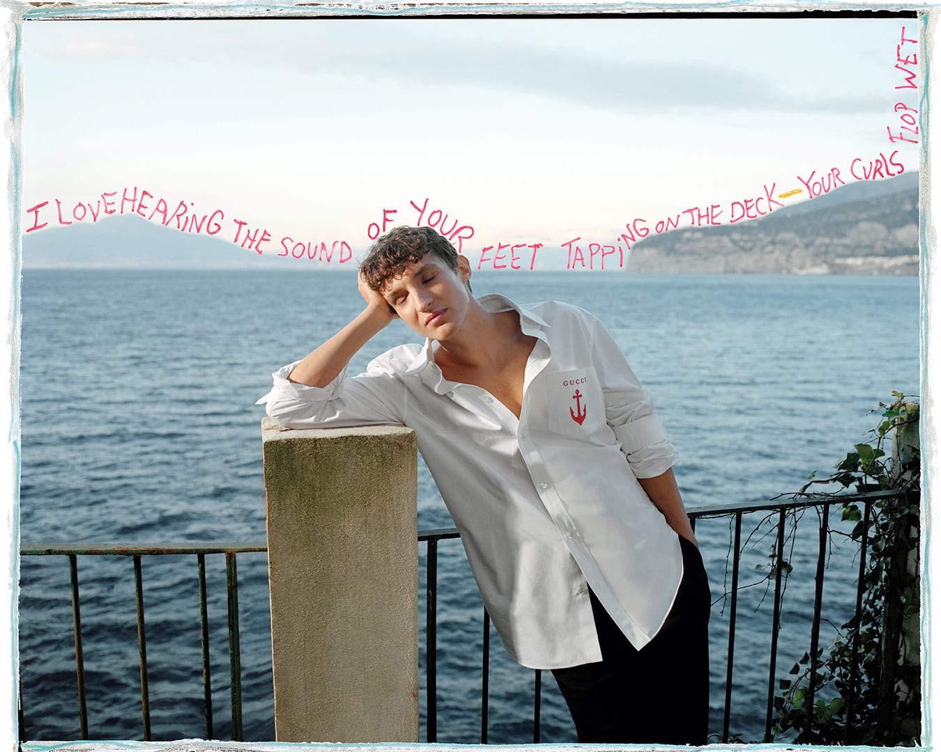Man wearing a white shirt leaning against a stone pillar by the ocean with handwritten text across the sky.