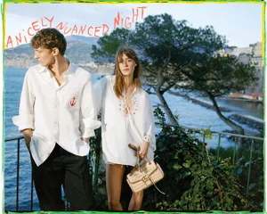 A man and woman in white shirts stand on a coastal balcony with water and a town in the background.