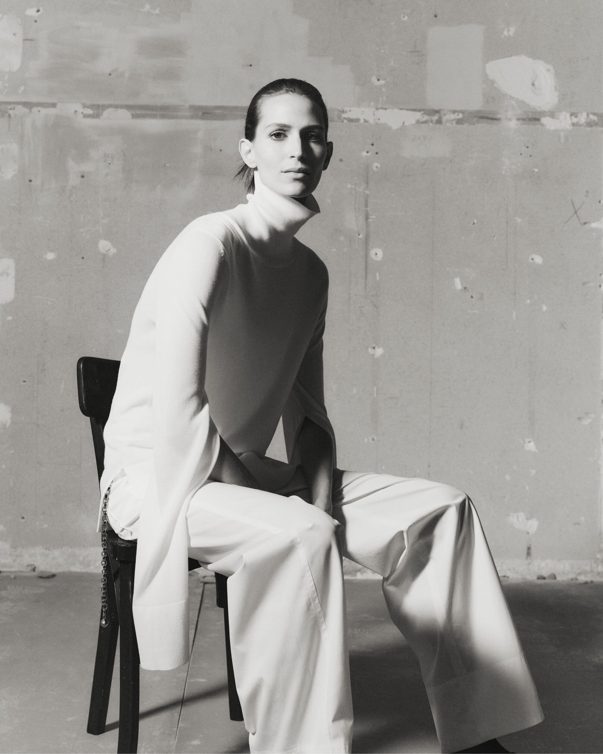 Jeanne Cadieu seated on a dark chair wearing a white turtleneck and trousers against a textured wall for Totême