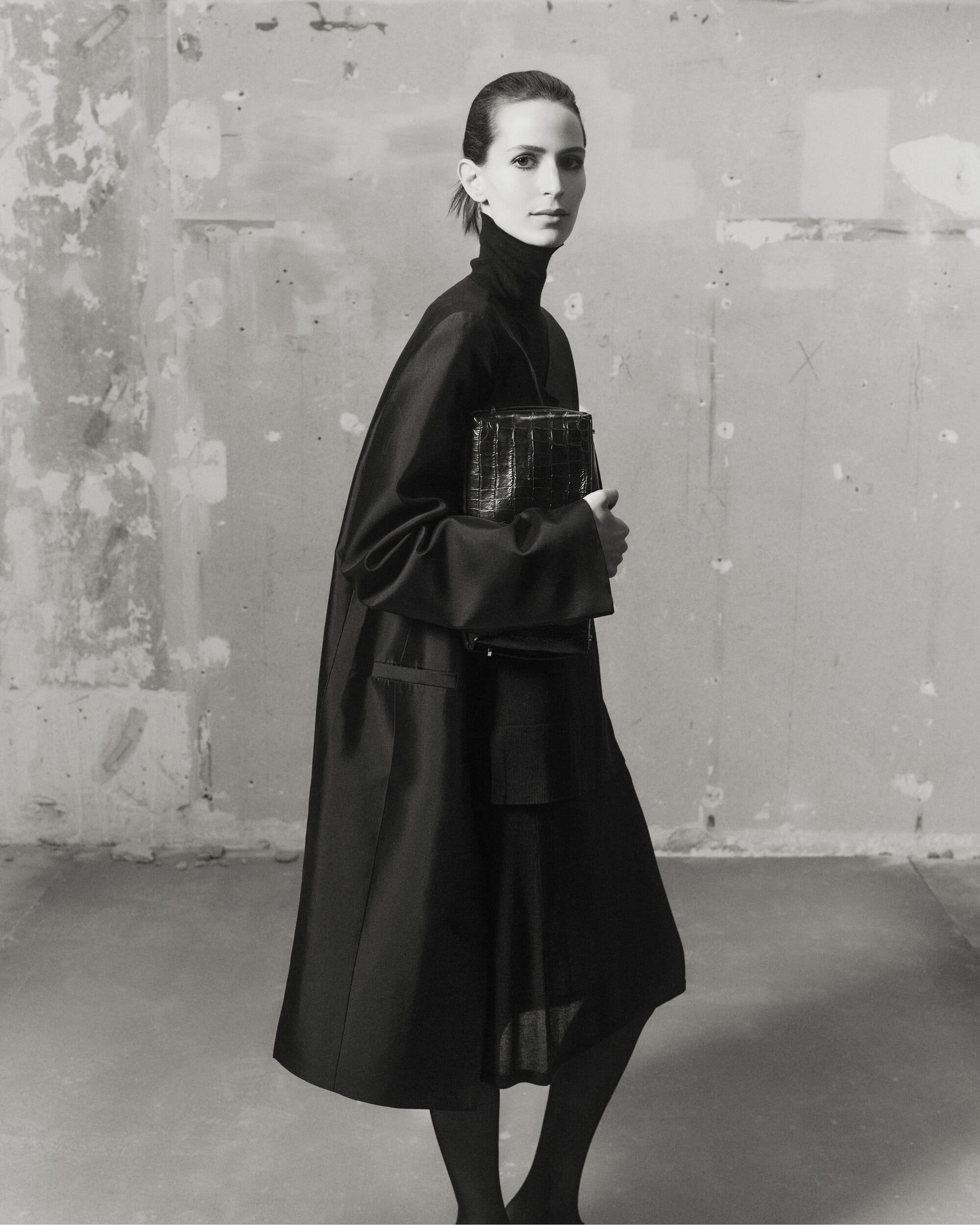 Model Jeanne Cadieu wearing a black turtleneck coat holding a dark handbag against a distressed concrete wall