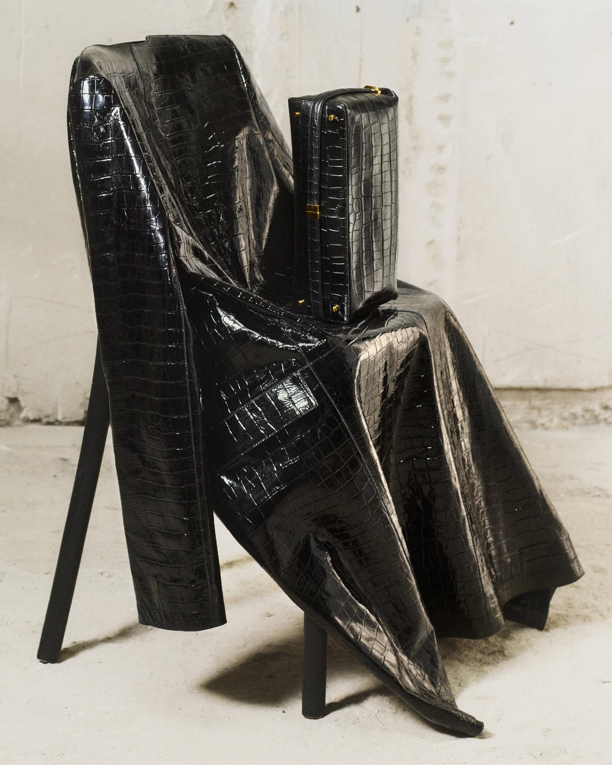 Black crocodile embossed leather coat draped over a chair with a matching upright handbag on a textured wall background