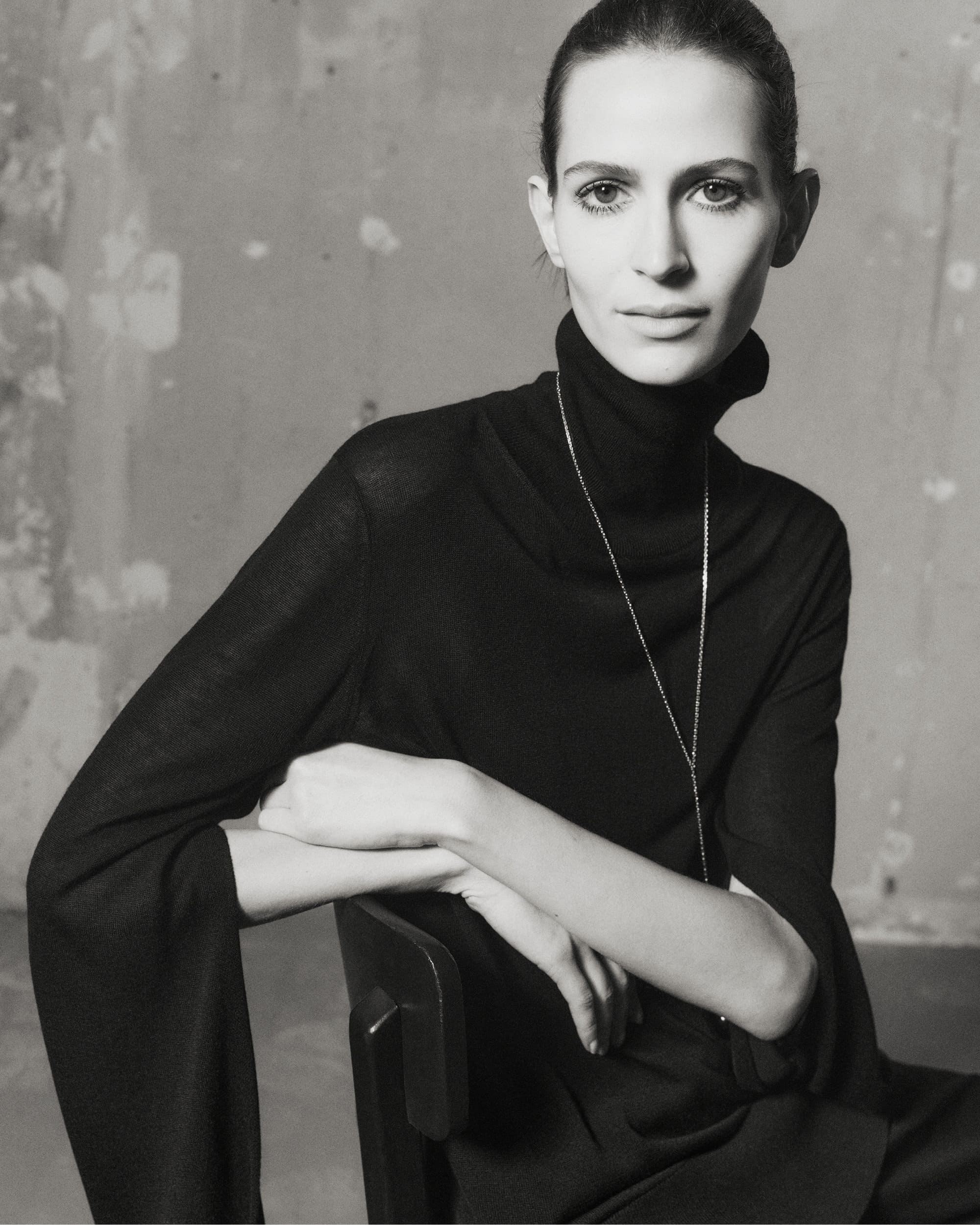Model Jeanne Cadieu wearing a black turtleneck sweater seated against a textured wall for Totême