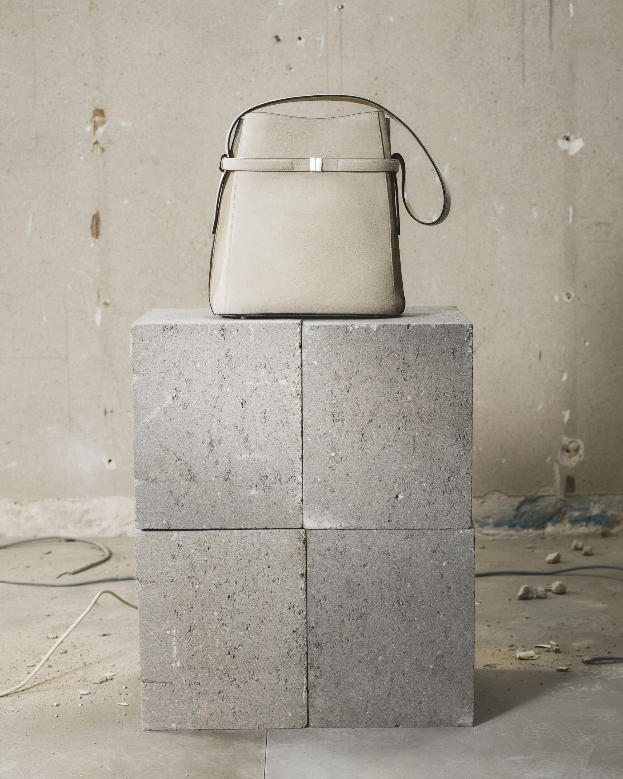 Beige leather bucket bag resting on a square concrete block against a weathered wall background