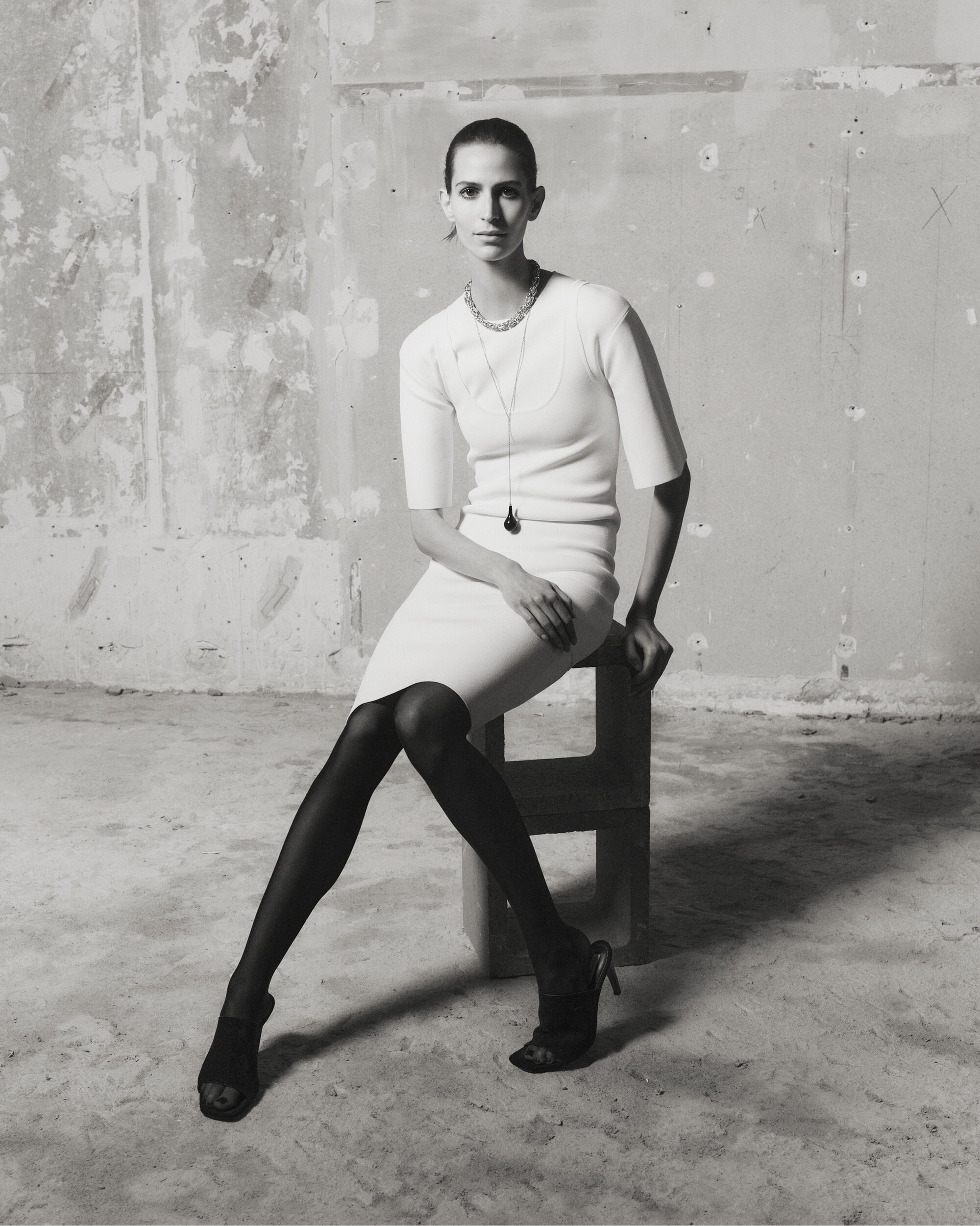 Model Jeanne Cadieu seated on a cinder block wearing a white ribbed dress and black tights for Toteme