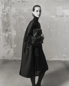 Model Jeanne Cadieu wearing a black turtleneck coat holding a dark handbag against a distressed concrete wall