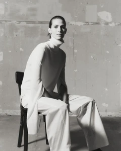 Jeanne Cadieu seated on a dark chair wearing a white turtleneck and trousers against a textured wall for Totême