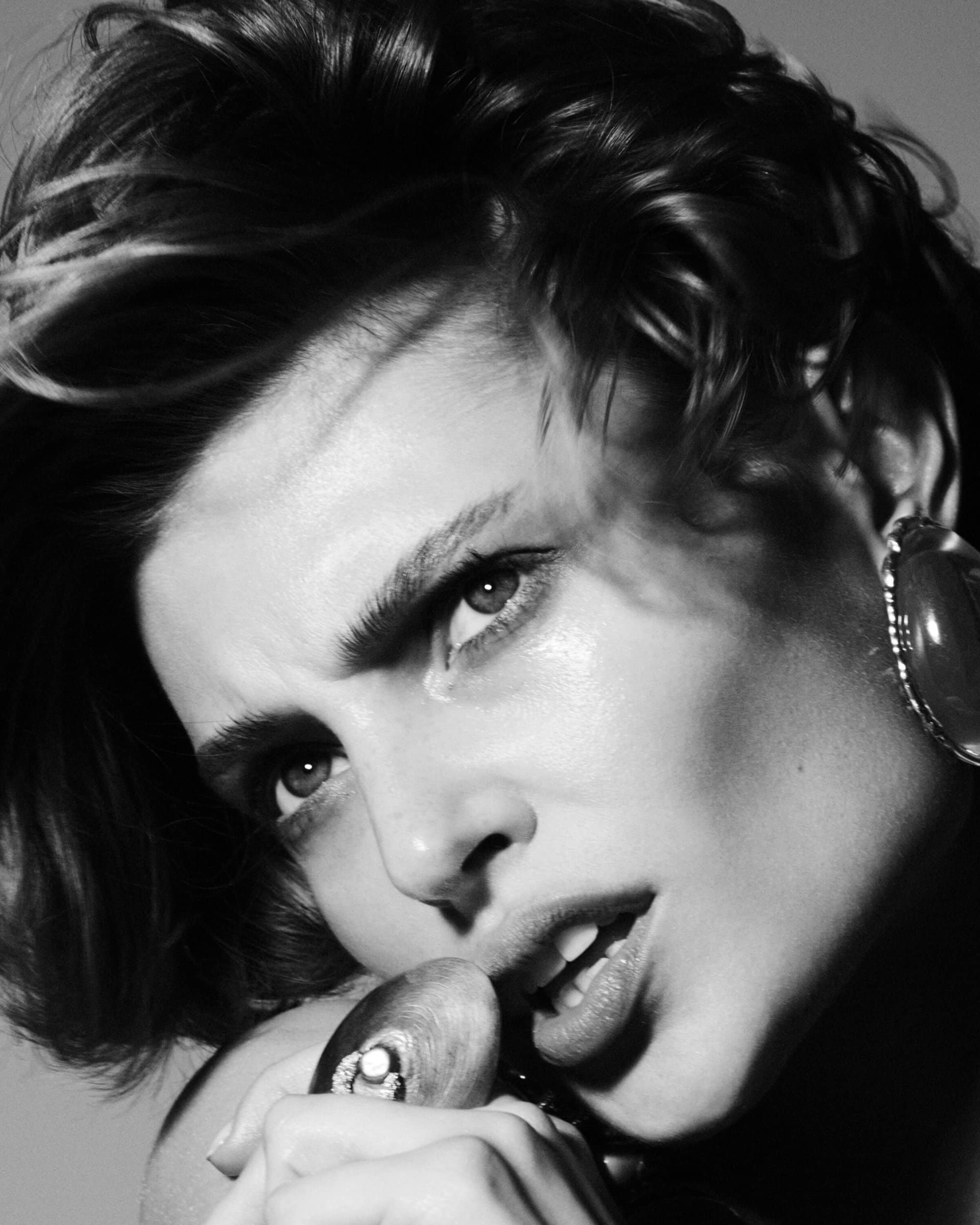 Close-up monochrome portrait of a woman wearing a large statement earring and holding a sculptural ring near her lips.