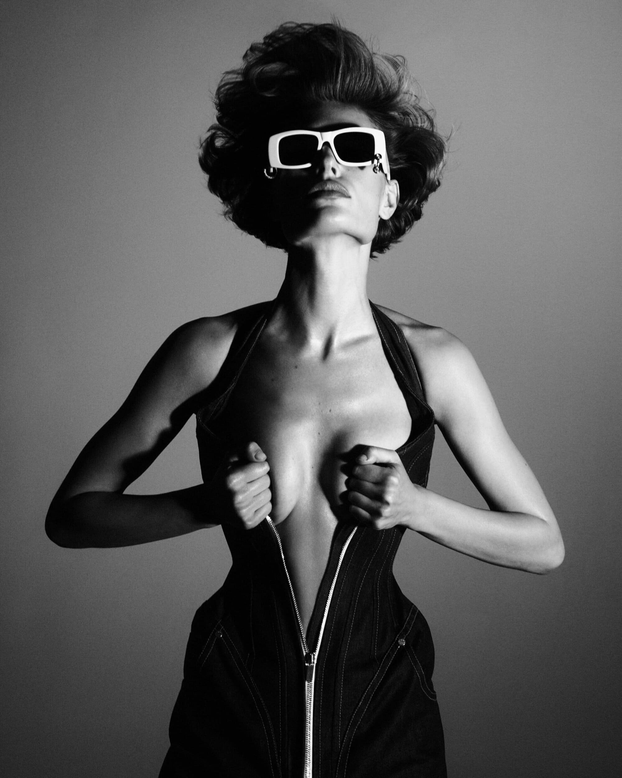 Model in white sunglasses unzipping dark fitted garment against neutral studio background
