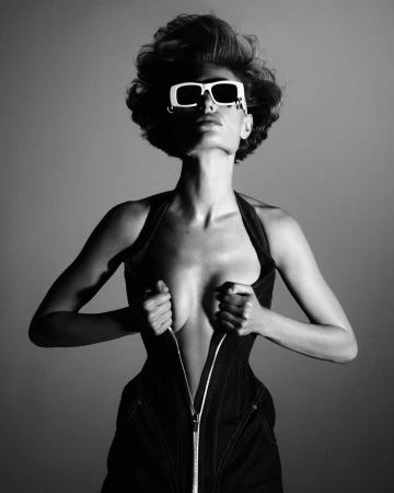Model in white sunglasses unzipping dark fitted garment against neutral studio background