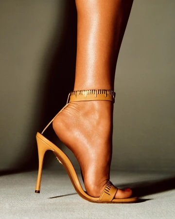Close-up of a leg in a tan high-heeled sandal with a gold measuring tape ankle strap against a grey seamless background