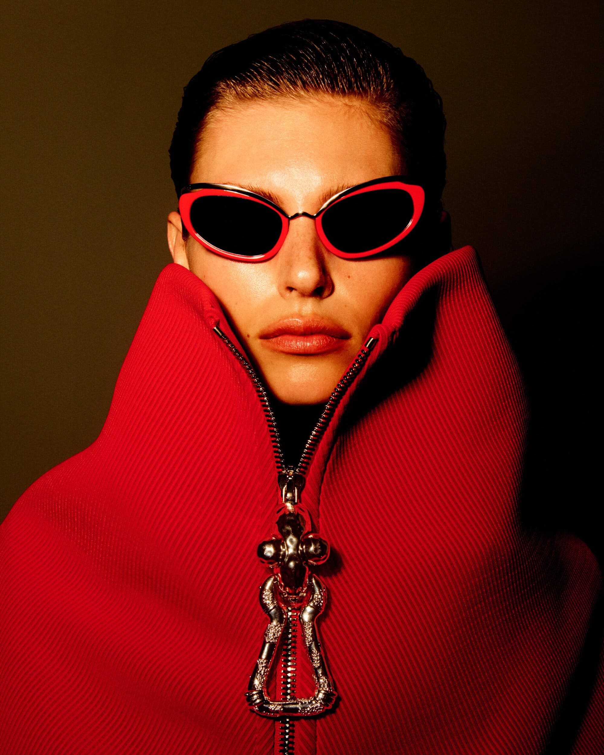 Model wearing red sunglasses and a high-collared red zipped garment with a metallic pendant pull by Schiaparelli.