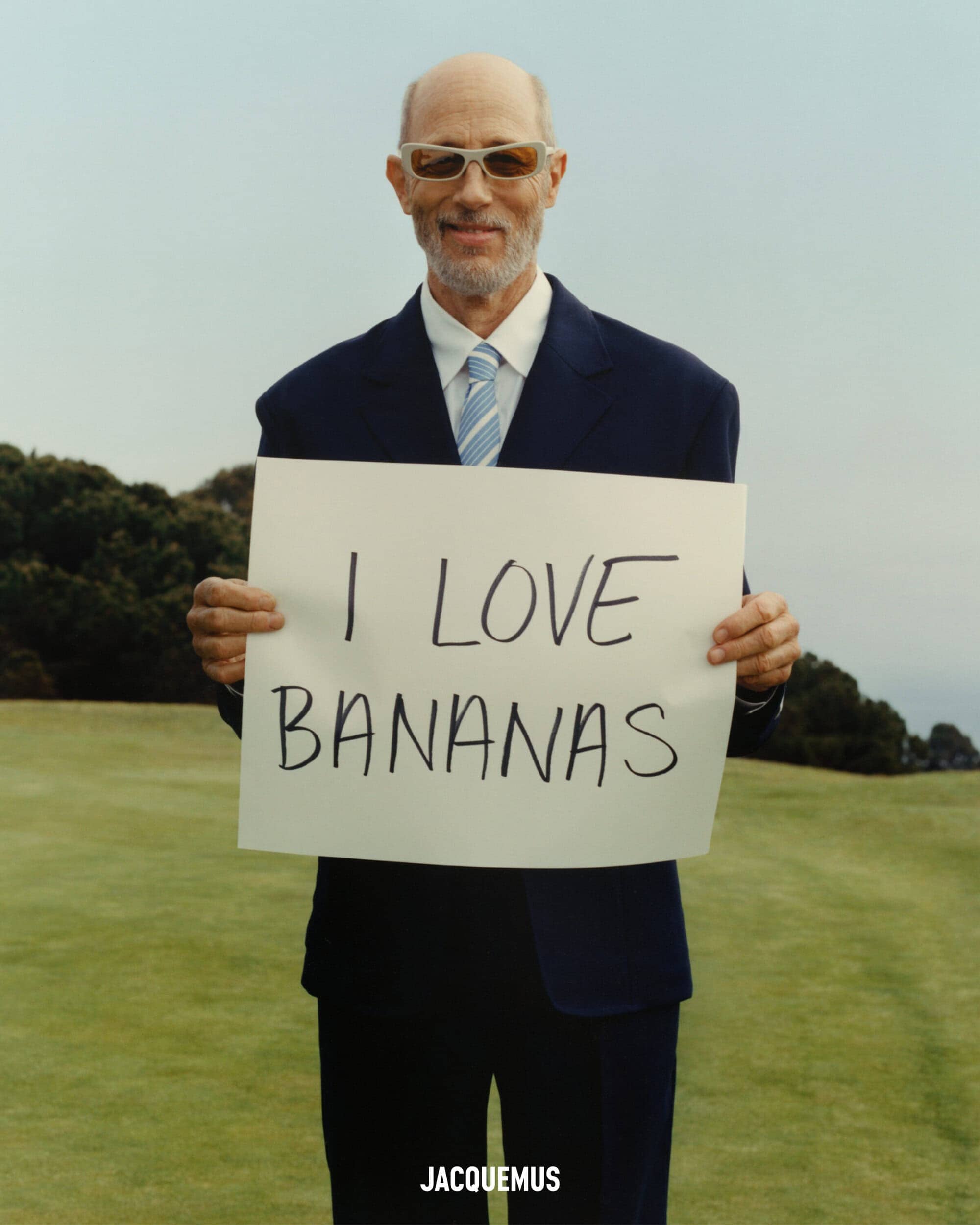 Jon Gries in a navy suit holding a white sign reading I LOVE BANANAS outdoors on grass