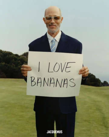Jon Gries in a navy suit holding a white sign reading I LOVE BANANAS outdoors on grass