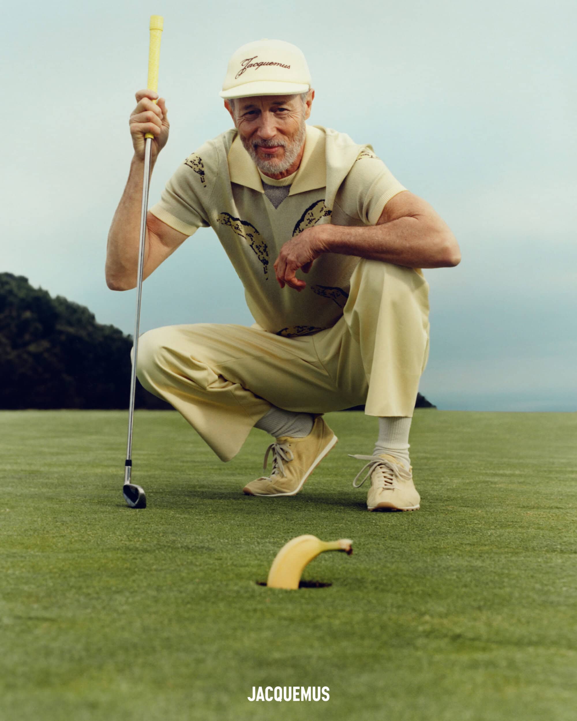 An older man crouches on a green golf course holding a club, looking toward a single banana sticking out of the hole in the ground.