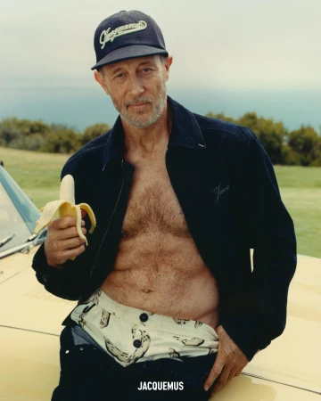 An older man wearing a dark cap, unbuttoned dark jacket, and patterned shorts holds a banana while standing outdoors.