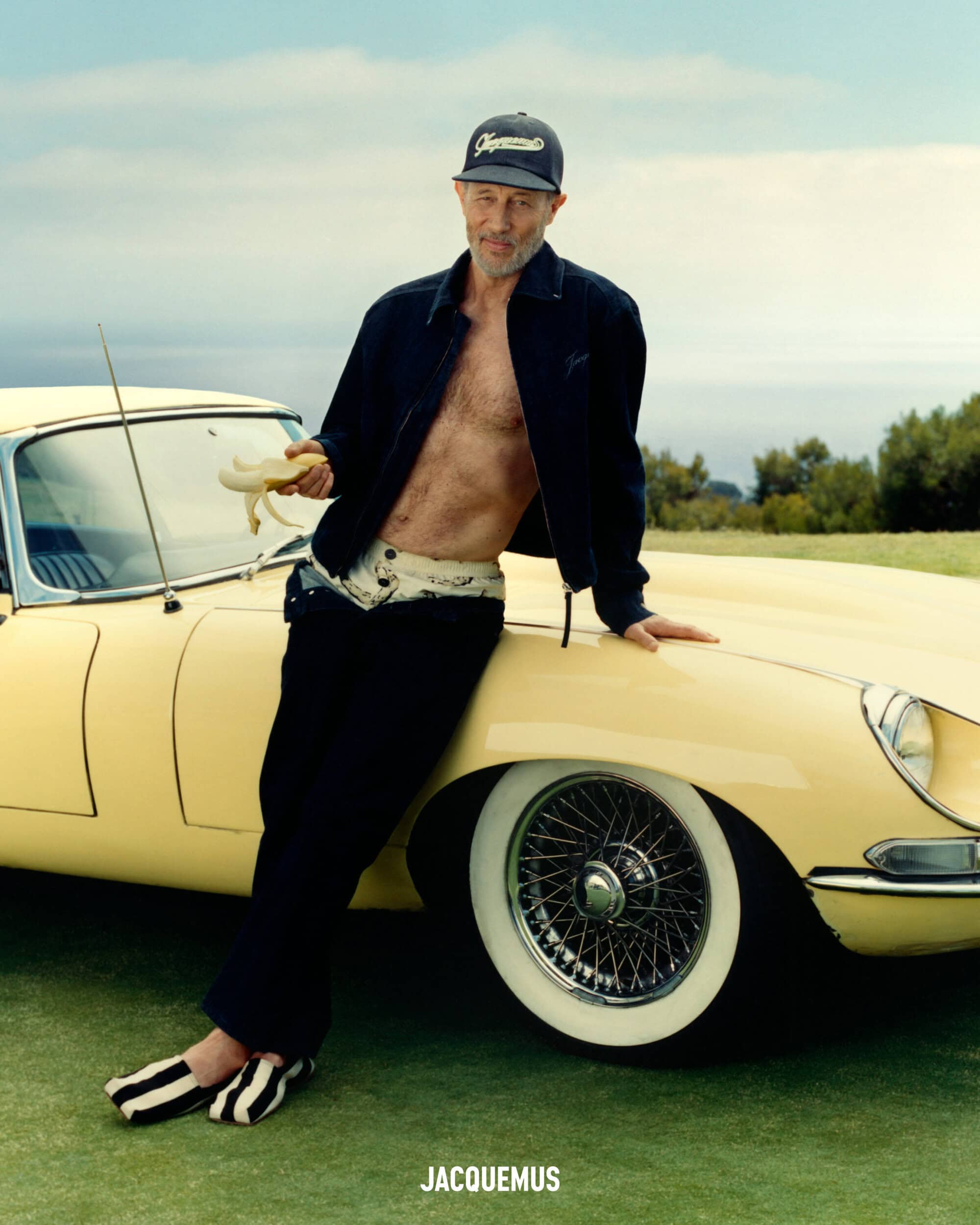 Jon Gries leaning against a yellow car wearing a dark jacket, baseball cap, and holding a banana for Jacquemus