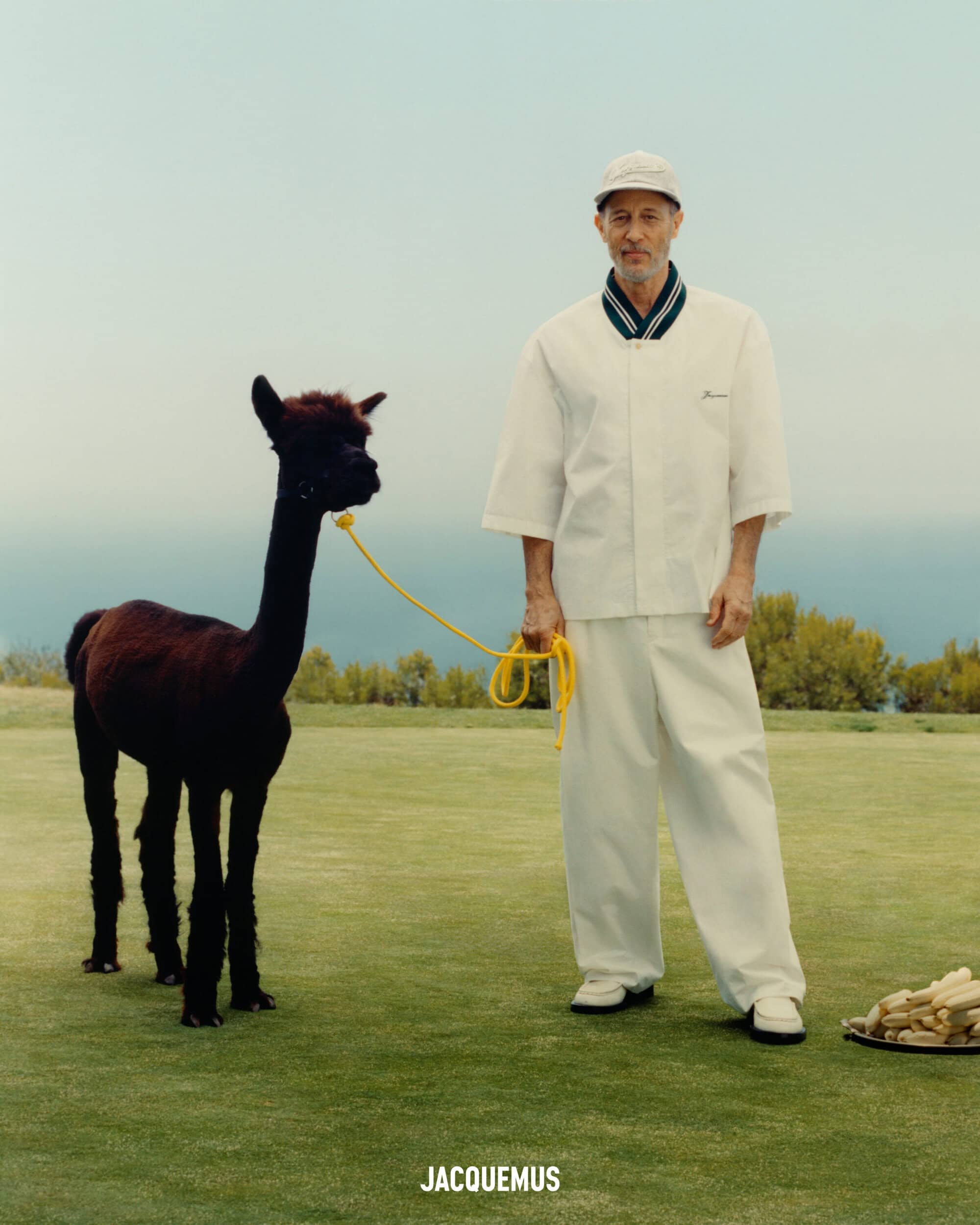 Jon Gries in a white outfit holding a yellow lead attached to a dark alpaca on a grassy field under a pale sky.
