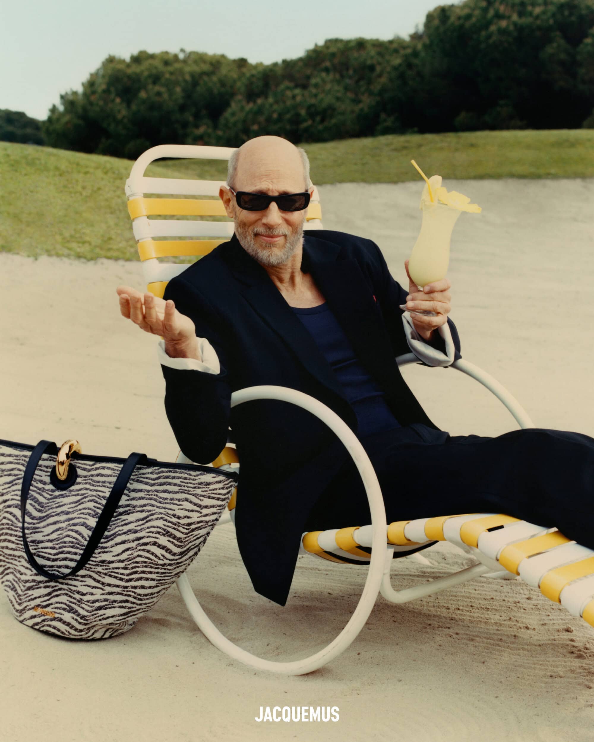 Jon Gries in dark sunglasses holding a cocktail, seated on a striped yellow lounger on sand for Jacquemus