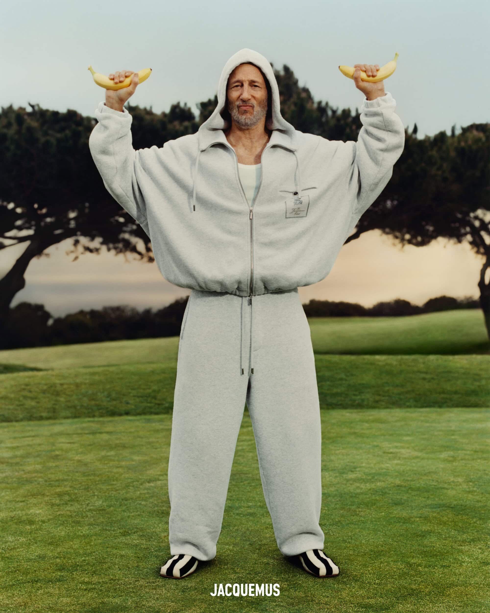 Jon Gries in a grey zip hoodie and sweatpants holding bananas up like weights outdoors on grass for Jacquemus