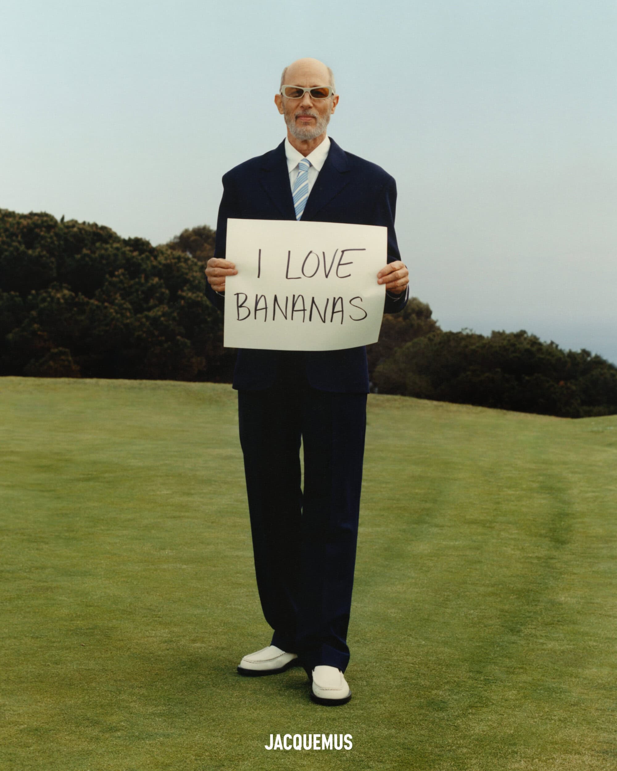 Jon Gries in a navy suit holding a white sign reading I LOVE BANANAS on a grassy hill for Jacquemus