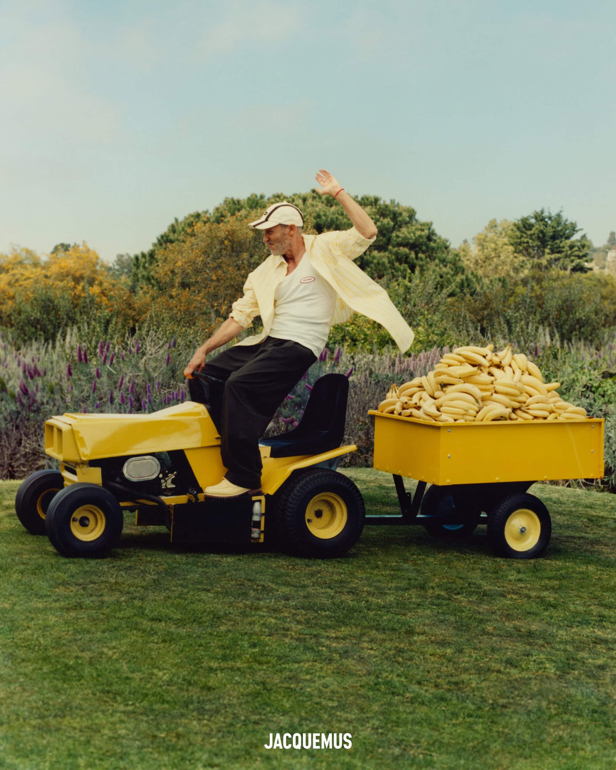 An older man waving while riding a yellow lawnmower pulling a cart filled with bananas across a grassy field.