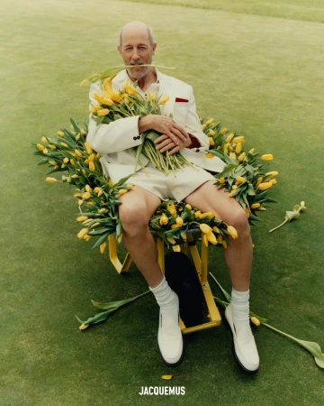 Jon Gries sitting in a yellow wheelbarrow filled with yellow tulips on a green lawn wearing a white suit