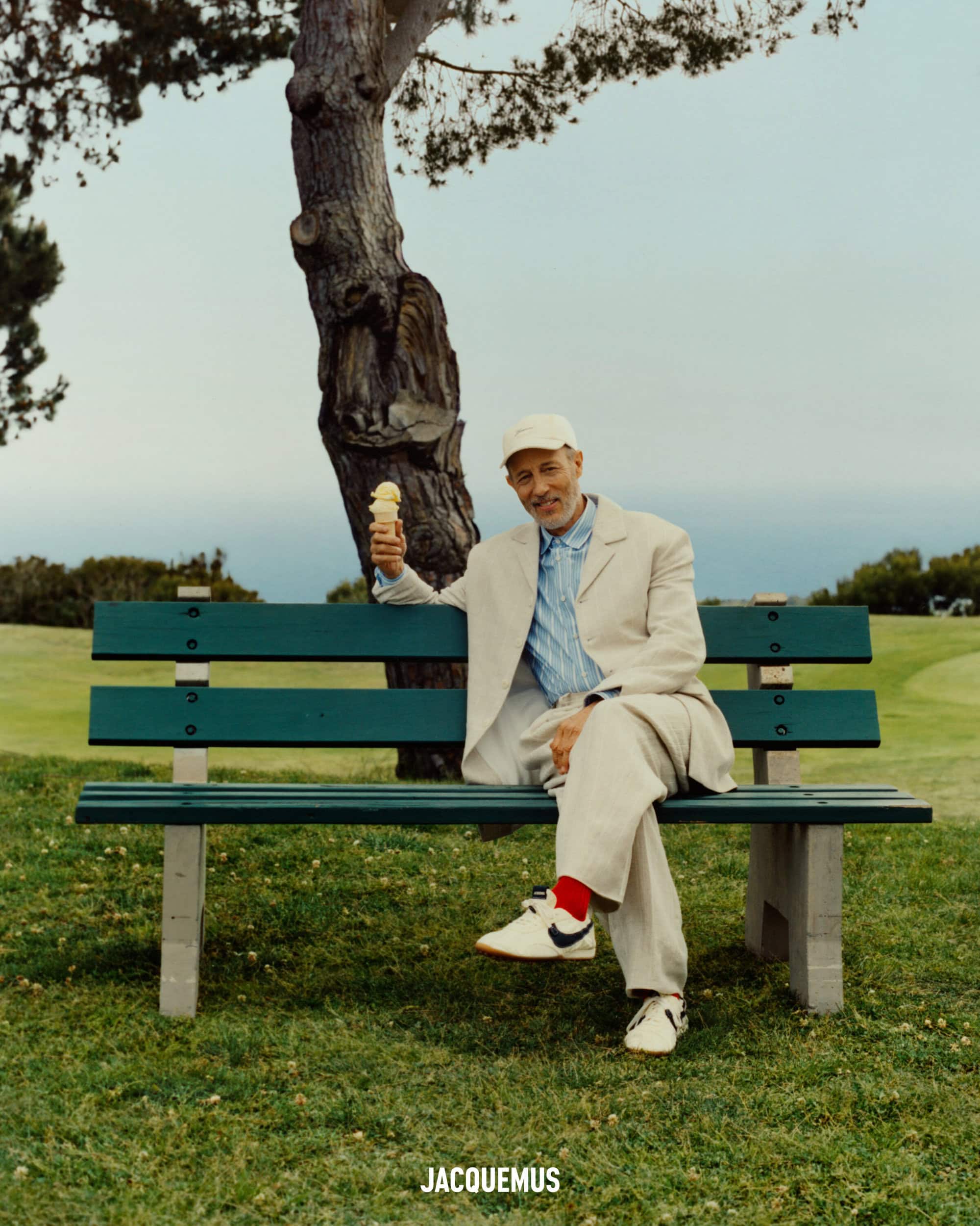 Jon Gries seated on a green park bench holding an ice cream cone for Jacquemus