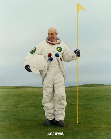 An older bald man wearing a white spacesuit holding a golf flag on a green grass field under an overcast sky.