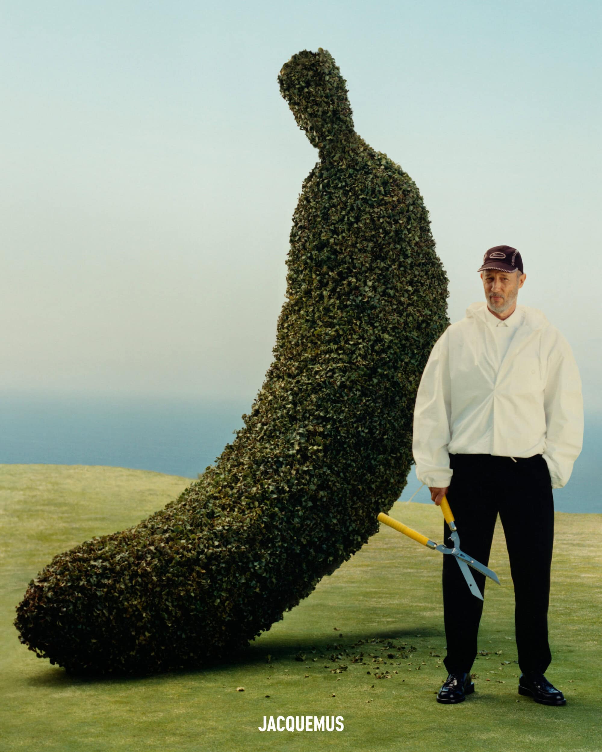 Man holding garden shears standing beside a giant banana-shaped topiary hedge on a green lawn