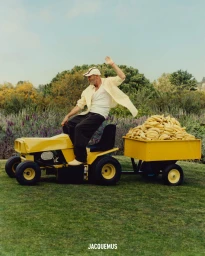 An older man waving while riding a yellow lawnmower pulling a cart filled with bananas across a grassy field.