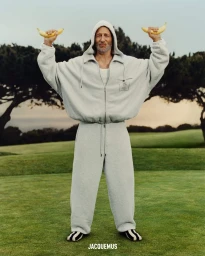 Jon Gries in a grey zip hoodie and sweatpants holding bananas up like weights outdoors on grass for Jacquemus