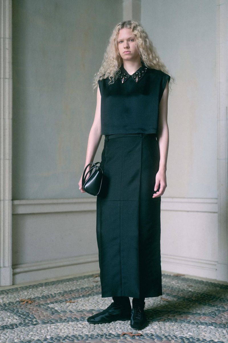 Blonde-haired person in a black two-piece outfit with a small black purse stands indoors on a stone-tiled floor near a wall with architectural details.