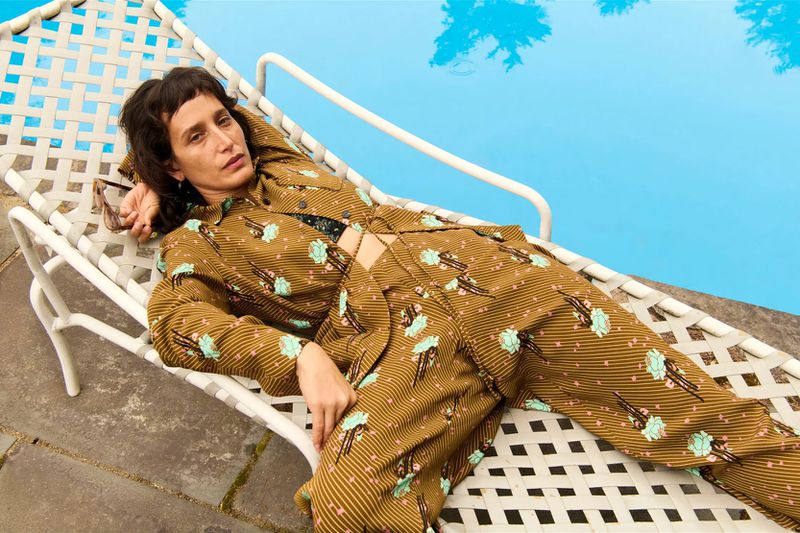 An adult female reclines on a white chaise lounge near a pool. She wears a patterned brown suit and dark sunglasses rest on the lounge.