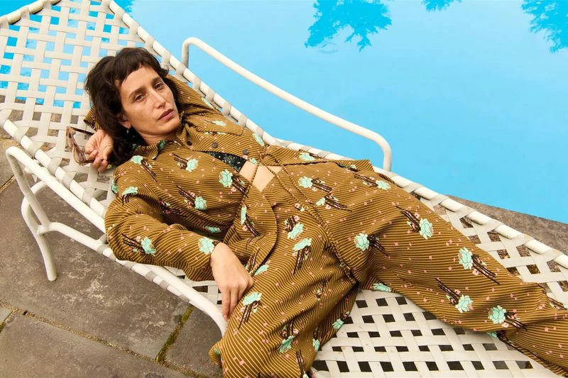 An adult female reclines on a white chaise lounge near a pool. She wears a patterned brown suit and dark sunglasses rest on the lounge.