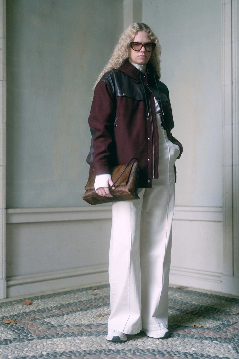 Full-length shot of a woman with blonde curly hair wearing a burgundy jacket, white pants, sunglasses and holding a brown purse.
