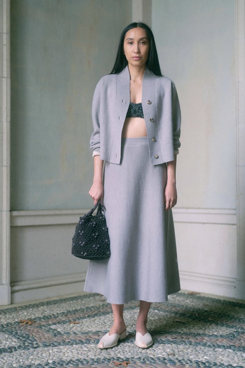 Elegant Asian woman poses in a grey skirt suit indoors, holding a knitted bag, complemented by neutral tones and a cobblestone floor.