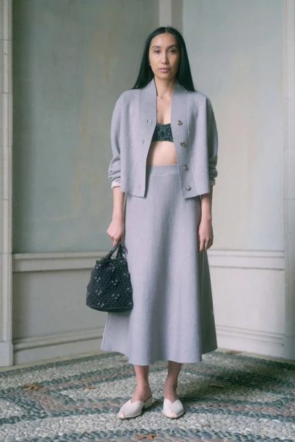 Elegant Asian woman poses in a grey skirt suit indoors, holding a knitted bag, complemented by neutral tones and a cobblestone floor.
