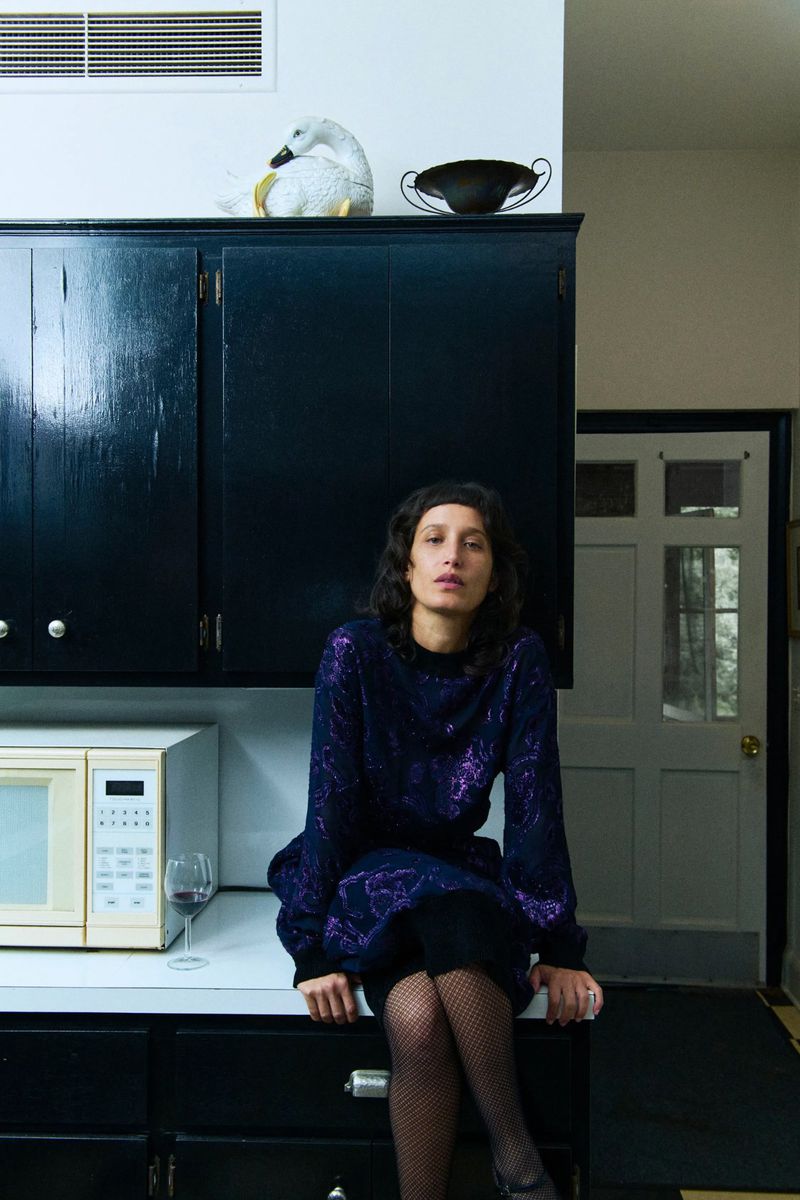 Indoor portrait of an adult woman sitting on a kitchen counter wearing a purple patterned long-sleeve dress and black pantyhose.