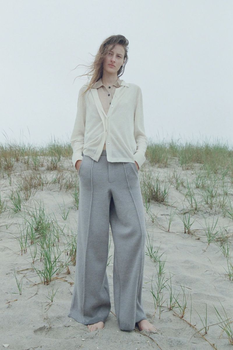 A woman in a light cardigan and grey pants stands on a sandy beach with dune grass against a neutral grey sky on an overcast day.