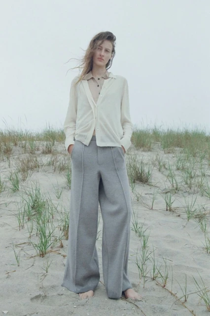 A woman in a light cardigan and grey pants stands on a sandy beach with dune grass against a neutral grey sky on an overcast day.