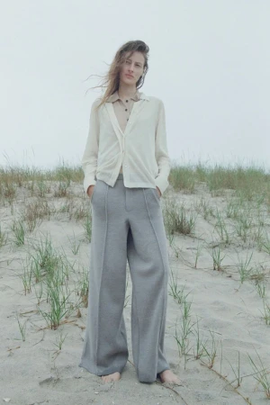 A woman in a light cardigan and grey pants stands on a sandy beach with dune grass against a neutral grey sky on an overcast day.