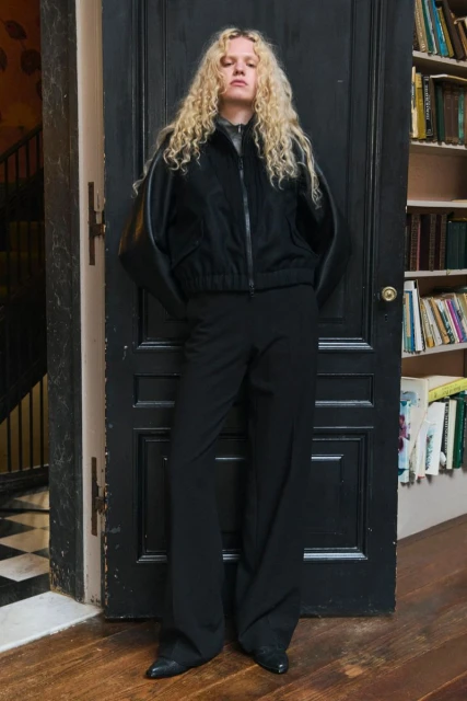 A curly-haired woman in a black jacket and wide-leg black pants stands in front of a dark wooden door with books in the background.
